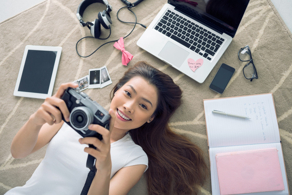 woman checking photos in camera while laying down alongsidde laptop and journals